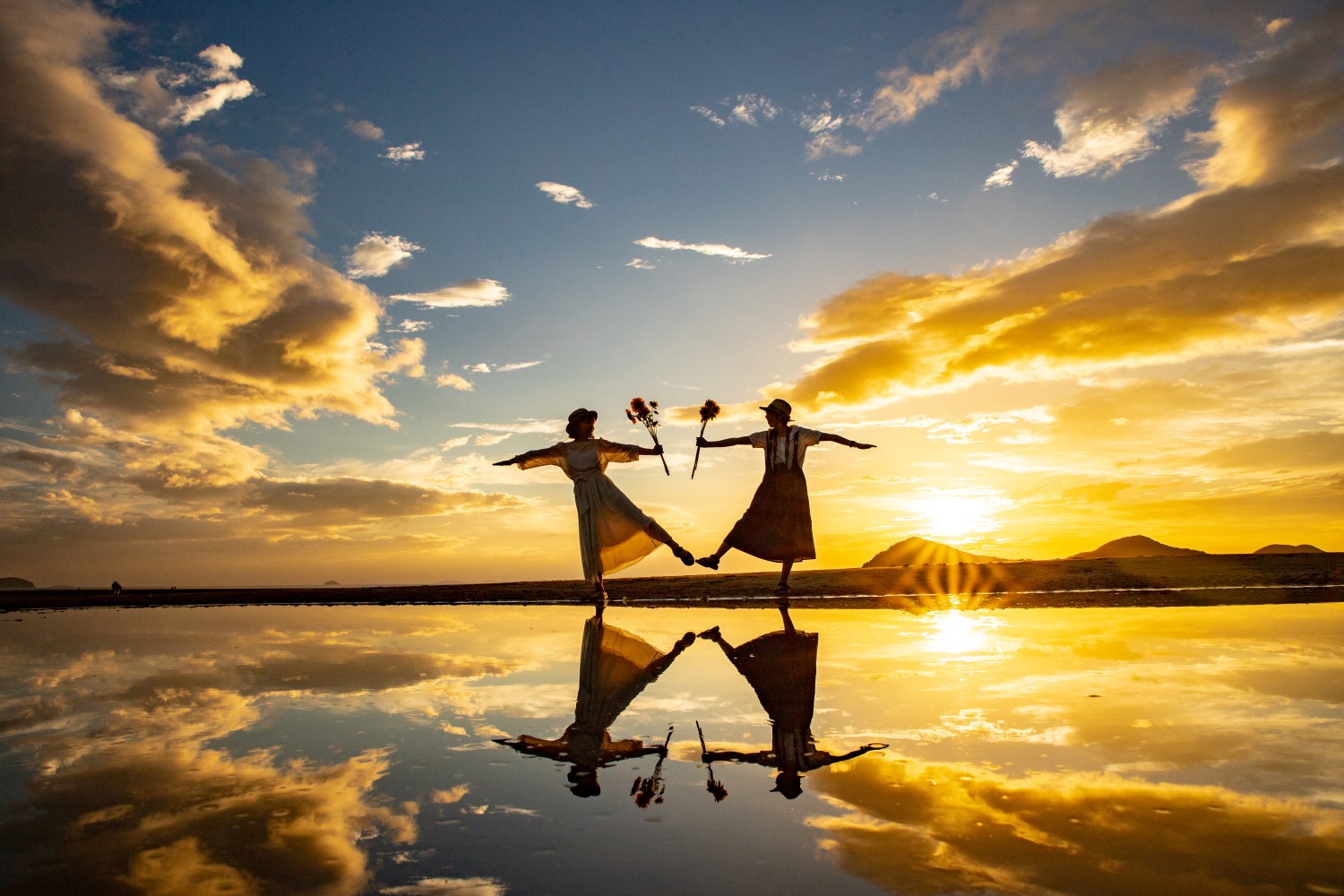 Two women standing on the beach reflected in the water below with the setting sun behind them. They are holding sunflowers and standing on one leg with the other extended, mirroring each other's pose.