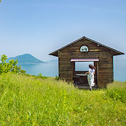 A small hut on Shishijima Island. A woman in a white dress is entering the hut. Behind it is a view of the Seto Inland Sea and islands.