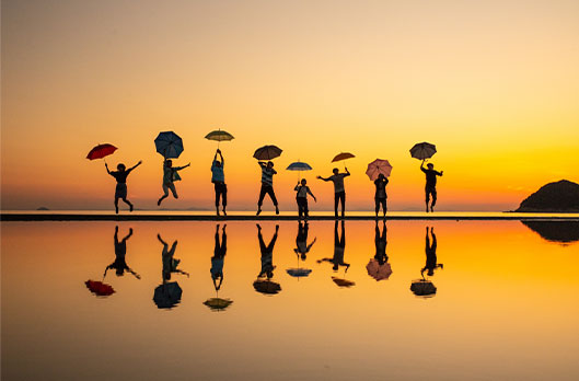 8 people lined on on Chichibugahama Beach. Each of them holds a colorful umbrella and is jumping at different heights. They are all reflected in the water below them with only a thin strip of land separating the water from the yellow sunset sky.