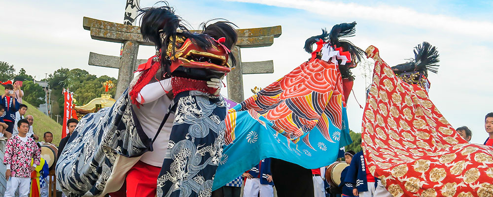 several people performing a lion dance in front of a torii gate. Many onlookers are visible on the sides.