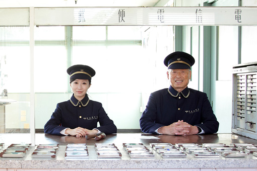 Kubota Saya, the artist who created the Missing Post Office, and Mr. Tanaka, the postmaster of the Missing Post Office, stand together at a counter. They are wearing matching post office uniforms.