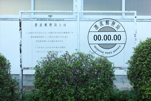 A sign in front of the Missing Post Office made of plexiglass in a clear frame. The left side has a description of the artwork in Japanese. The right side has a large emblem which looks like a post stamp and says 'Missing Post Office' in English and Japanese.