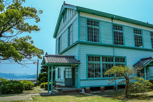 The Awashima Maritime Museum as seen from behind. The ocean is visible in front of the teal, 2-story wooden building. Many large windows dot the walls.