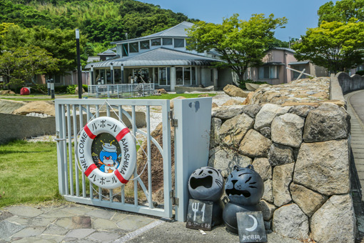 The gate to Le Port Awashima, set into a stone wall. A life ring hangs on the gate with 'welcome,' 'Le Port', and 'Awashima' written on it. Le Port Awashima is visible in the background, a rounded 2-story building with many windows.