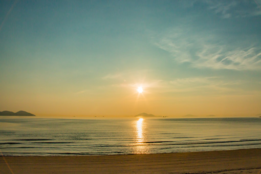 A wide, empty beach with islands visible in the distance across a calm sea. The sunset is perfectly centered in frame.