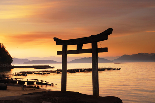 The torii gate at sunset. The gate is located at the edge of the water and is silhouetted against the yellows and purples of the sunset. Fishing equipment is visible behind the gate.