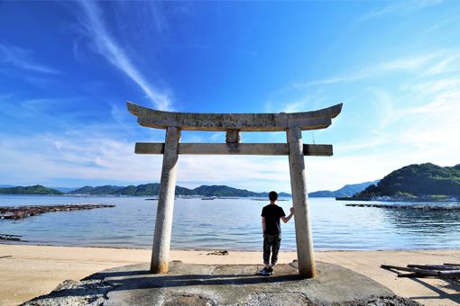 The torii gate during the day. A man leans against the inner right side of the gate facing the sea, which has reached almost to the base of the gate. The sky and sea are both a clear blue.