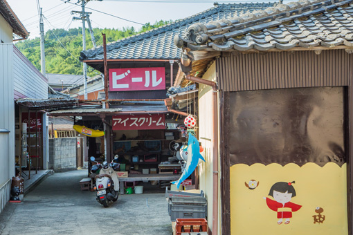 The outside of Takeuchi General Store. It is a Japanese-style building set into an alley with many goods visible and large signs saying 'beer' and 'ice cream' in Japanese.