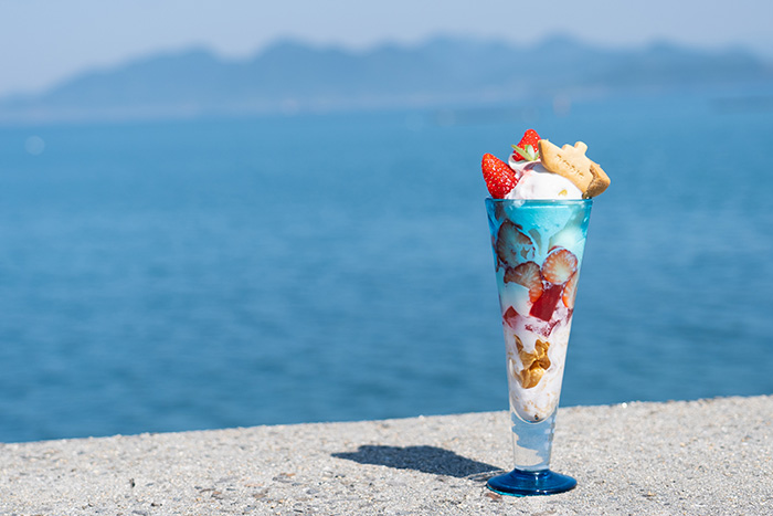 A parfait with strawberries and a cookie shaped like a boat is on a seawall in front of a very blue ocean.
