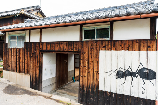 The outside of Art Canvas Awashima, a Japanese-style home with a charred wood and plaster facade. A large painting of a black ant on a white background is painted next to the entrance.