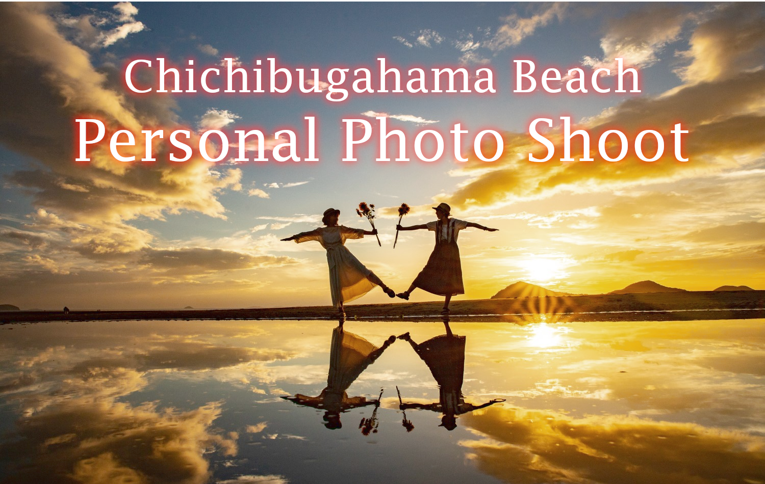 Two women standing on the beach reflected in the water below with the setting sun behind them. They are holding sunflowers and standing on one leg with the other extended, mirroring each other's pose.