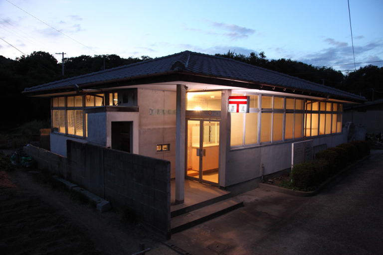 The outside of the Missing Post Office, a square, concrete building with many windows. There is a sign above the door which prominently features Japan's symbol for post.